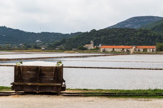 Vintage wooden harvest cart on rails at Ston salt pans with shallow evaporation ponds and historic stone building behind, Croatia