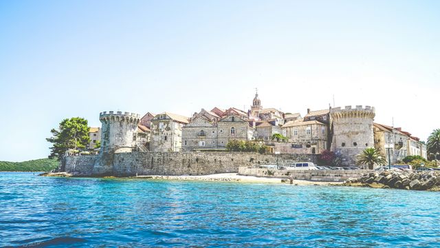 Ancient walled coastal town with stone buildings overlooking crystal-clear turquoise waters of the Adriatic Sea.